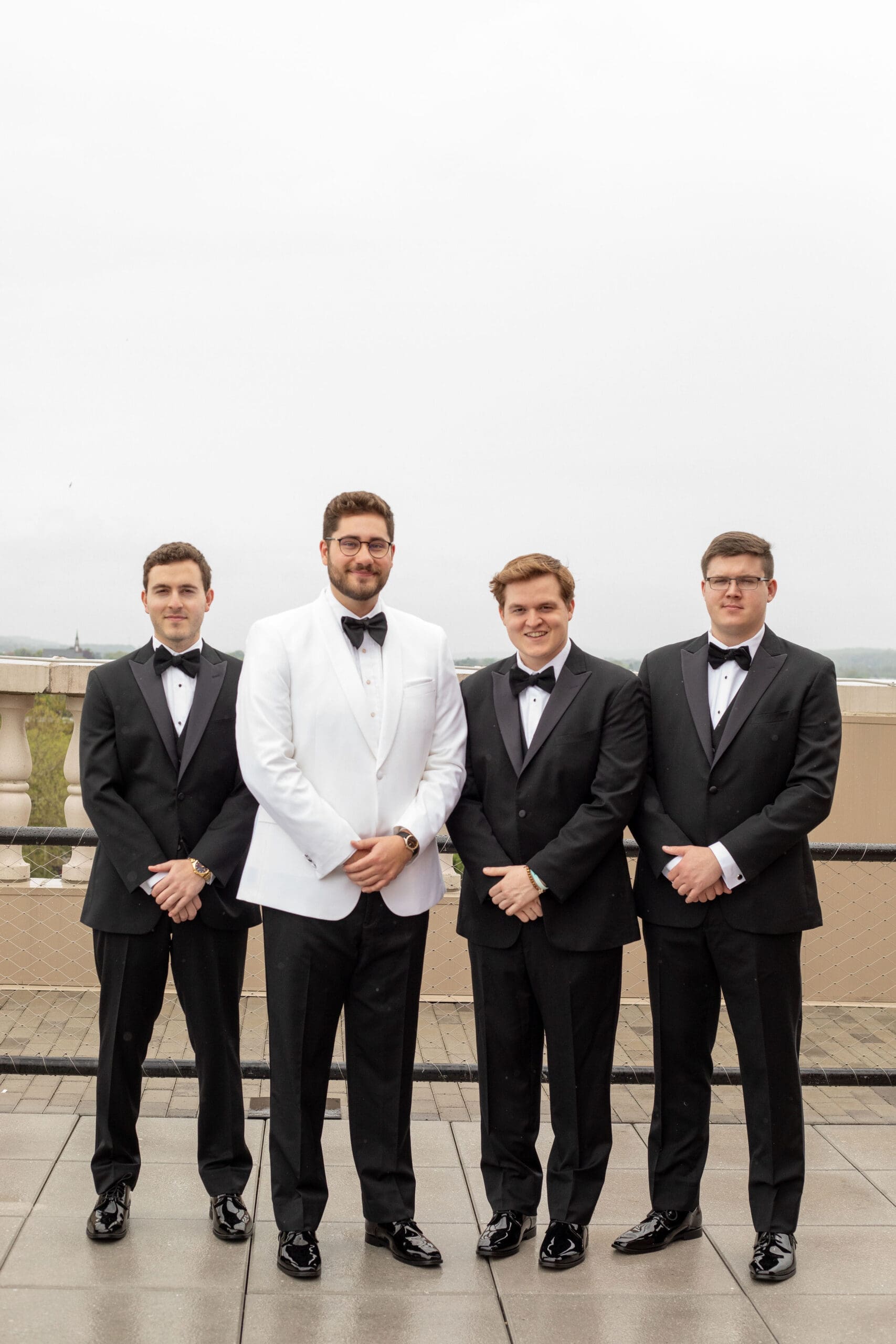 Groom and groomsmen on an overcast day on a rooftop balcony. The groomsmen are wearing black tuxedos, while the groom has a white tuxedo jacket 